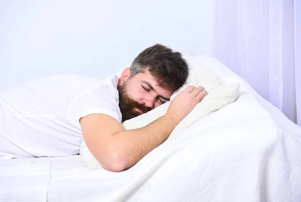 Hombre de camisa acostado en la cama, pared blanca en el fondo. Un tipo con la cara tranquila durmiendo en sábanas blancas y almohada. Macho con barba y bigote durmiendo, relajándose, durmiendo la siesta, descansando. Concepto siesta y siesta — Foto de Stock