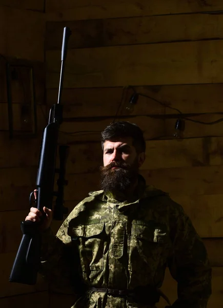 Hombre con barba lleva ropa de camuflaje, fondo interior de madera. Macho en cara estricta en la casa de los guardabosques. Hunter, brutal hipster con arma lista para cazar. Concepto de cazador —  Fotos de Stock