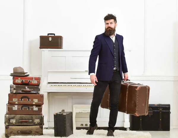Luggage and vacation concept. Man, traveller with beard and mustache with luggage, luxury white interior background. Macho elegant on strict face stands near pile of vintage suitcase, holds suitcase
