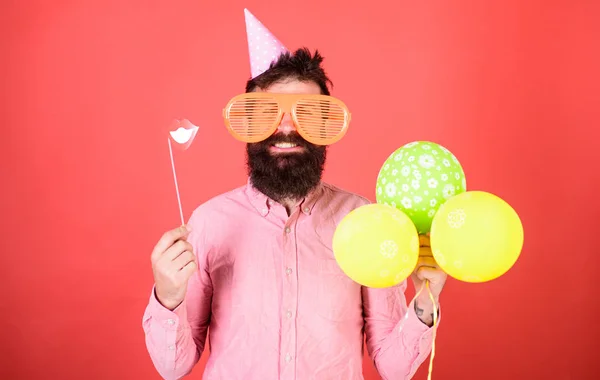 Skäggig man med part tillbehör, överraskning koncept. Man med papper läppar, enorma galen glasögon och ballonger isolerad på röd bakgrund. Hipster med långt skägg bär Rosa skjorta och födelsedag cap — Stockfoto