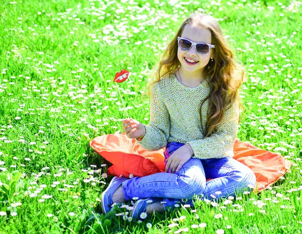 草地、緑の背景に芝生の上に女の子が座っています。子供サングラス段ボール草原でのフォト セッションの唇を笑顔でポーズします。陽気な顔の女の子は、アウトドア レジャーをお過ごしください。ファッショニスタのコンセプト — ストック写真