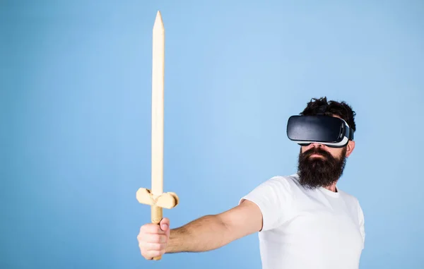 Gamer concept. Guy with head mounted display holds sword, play fighting game in VR. Man with beard in VR glasses, light blue background. Hipster on serious face enjoy play game in virtual reality