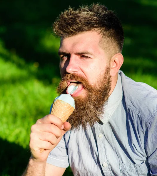 Conceito de delicadeza. Homem com barba longa come sorvete, enquanto se senta na grama. Homem barbudo com cone de gelado. Homem com barba e bigode no rosto sexy lambe sorvete, grama no fundo, desfocado — Fotografia de Stock