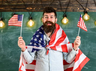 Vatansever eğitim konsepti. Öğrenci değişim programı. Amerikan öğretmeni Amerikan bayraklarıyla el sallıyor. Mutlu yüzlü sakallı ve bıyıklı bir adam ABD bayrakları taşıyor, sınıfta, arka planda karatahta.