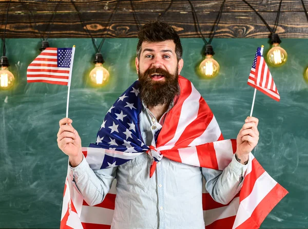 Patriottisch onderwijsconcept. Studenten uitwisselingsprogramma. Amerikaanse leraar zwaait met Amerikaanse vlaggen. Man met baard en snor op vrolijk gezicht draagt vlaggen van de VS, in de klas, schoolbord op achtergrond — Stockfoto
