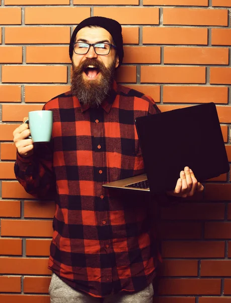 Bearded brutal caucasian hipster holding laptop with cup — Stock Photo, Image