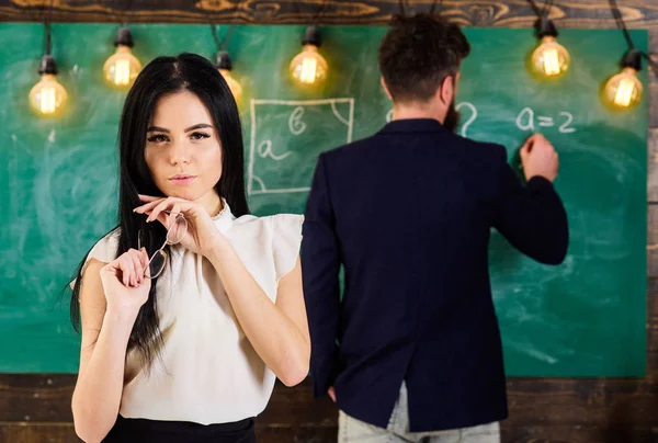 Meisje student spelen met brillen terwijl leraar op het schoolbord schrijft. Lady op dromerige gezicht niet aandacht besteden aan leraar en les. Student met lange haren in formele slijtage ziet er aantrekkelijk en sexy — Stockfoto