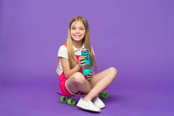 Chica en la cara sonriente sostiene con botella de agua mientras se sienta en el monopatín, fondo violeta. Niña con el pelo largo bebe agua. Concepto de balance hídrico. Chica se preocupa por la salud y el equilibrio hídrico —  Fotos de Stock