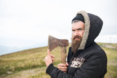 Sıkı yüzündeki sakallı hippi keskin balta, manzarası arka plan, kopya alanı üzerinde tutar. Acımasız ve sakallı oduncu balta tutar. Acımasız oduncu kavramı. Şapka ve sıcak ceketli adamı vahşice görünüyor