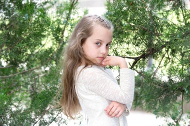 Yaz mevsiminde yeni bir cilde sahip çocuk manken. Moda elbiseli kız yeşil ağaçta poz veriyor. Açık havada uzun sarı saçlı küçük bir çocuk. Moda güzelliği ve saç stili. Cilt bakımı ve tazeliği