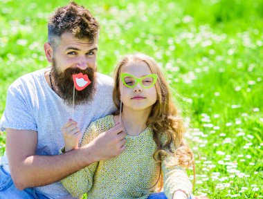 Baba ve kızı, grassplot, arka planı yeşil çimenlerin üzerinde oturur. Aile açık havada eğlence harcamak. Çocuk ve babası eyeglases ve dudakları ile poz booth öznitelikleri fotoğraf. Babalık kavramı