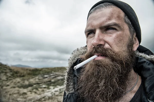 Hombre con barba larga y bigote fumando cigarrillo. Concepto de brutalidad. Hipster en la cara estricta con barba se ve brutalmente durante el senderismo y fumar. Hombre con apariencia barbuda brutal, desordenado y sin afeitar —  Fotos de Stock