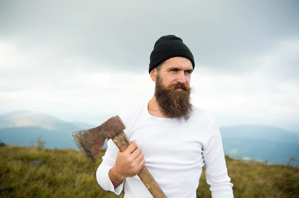 Hipster con barba en la cara estricta sostiene hacha, horizonte en el fondo. Lumberjack brutal y barbudo sostiene hacha mientras está parado en la cima de la montaña. Concepto de senderismo. Hipster se ve brutal y elegante con hacha — Foto de Stock