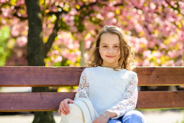 背景には デフォーカスの桜の木のベンチに座っている笑顔の女の子 概念を説明します バックパックでかわいい子では 日当たりの良い春の日をお楽しみください 桜の近くの公園を散歩中の女の子 — ストック写真