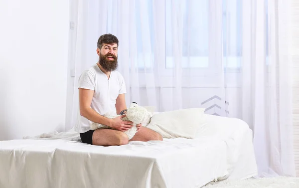 Um tipo a sorrir a brincar com um urso de peluche. Macho com barba e bigode relaxante com brinquedo de pelúcia após a sesta. Homem senta-se na cama e segura brinquedo, cortinas brancas no fundo. Conceito de memórias infantis — Fotografia de Stock