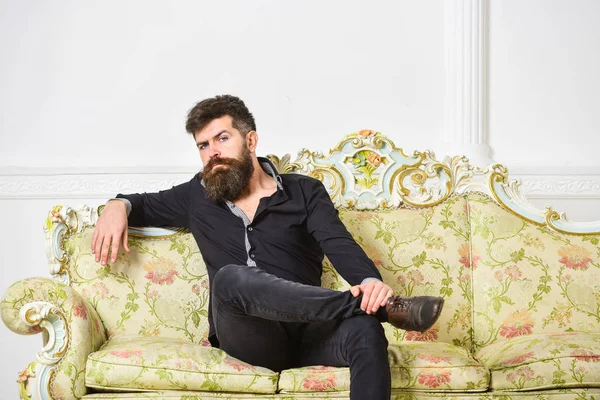 Hombre con barba y bigote pasa el ocio en la sala de estar de lujo. Hipster en cara pensativa se sienta solo. Propietario de apartamento de lujo se sienta en el sofá, fondo blanco de la pared. Concepto rico y solitario — Foto de Stock