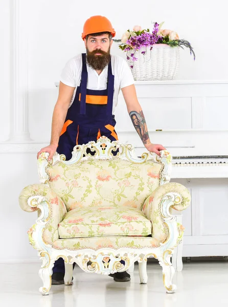 Delivery service concept. Courier delivers furniture in case of move out, relocation. Man with beard, worker in overalls and helmet lean on armchair, white background. Loader carries armchair