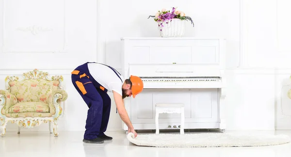 Relocating concept. Loader wrappes carpet into roll. Man with beard, worker in overalls and helmet rolling carpet, white background. Courier delivers furniture in case of move out, relocation