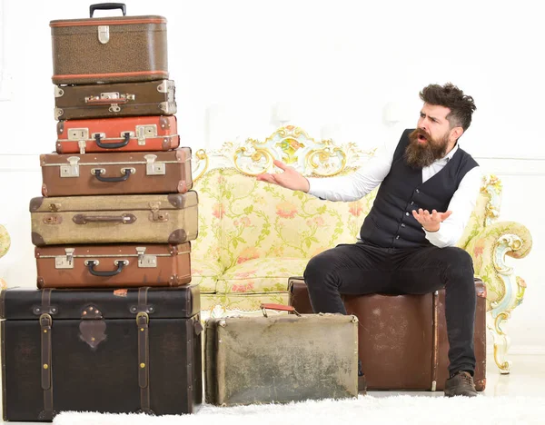 Macho elegant on surprised face sits shocked near pile of vintage suitcase. Luggage and travelling concept. Man, butler with beard and mustache delivers luggage, luxury white interior background