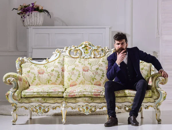 Conceito de moda e estilo. Macho atraente e elegante no rosto sério e expressão atenciosa. Homem com barba e bigode vestindo terno clássico elegante, senta-se no sofá à moda antiga ou sofá — Fotografia de Stock