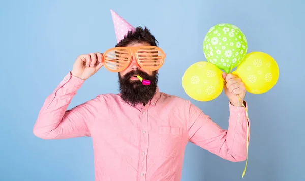 Hipster mit buschigem Bart feiert Geburtstag. Bärtiger Mann posiert in Geburtstagskappe mit riesiger Brille und bunten Luftballons auf blauem Hintergrund. Mann mit gestutztem Bart bläst Party-Pfeife — Stockfoto