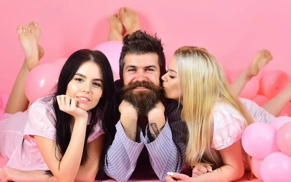Man with beard and mustache attracts blonde and brunette girls. Threesome on smiling faces lay near balloons. Girls fall in love with bearded macho, pink background. Alpha male concept — Stock Photo, Image