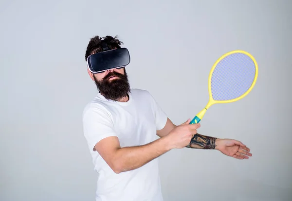 Guy with VR glasses play tennis with racket and ball. Man with beard in VR glasses beating pitch, grey background. Virtual sport concept. Hipster on busy face use modern technology for sport games