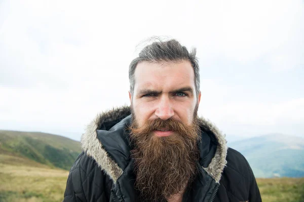 Man with long beard and mustache wears jacket. Hipster on strict face with beard looks brutally while hiking. Hermit concept. Man with brutal bearded appearance, brutal unshaven man looks untidy — Stock Photo, Image