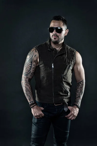 Fashion macho in trendy sunglasses. Tattoo model with beard on unshaven face. Bearded man with tattoo on strong arms. Tattooed man with biceps and triceps. Fashion style and trend, vintage filter