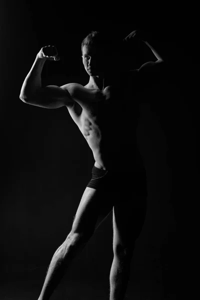 Macho met gespierd lichaam die zich voordeed als bodybuilder, zwarte achtergrond. Bodybuilding concept. Bodybuilder in zwarte onderbroeken toont zijn spieren. Silhouet van atleet demonstreren gespierd lichaam — Stockfoto