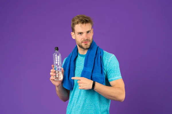 El deportista señala con el dedo la botella de plástico sobre fondo violeta. El hombre sostiene la botella de agua. Atleta en camiseta azul y pantalones cortos con toalla después del entrenamiento. Sed y deshidratación. Actividad deportiva y energía — Foto de Stock