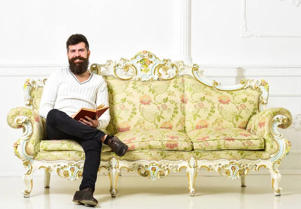 Mann mit Bart und Schnurrbart sitzt auf barockem Sofa, hält Buch, weißer Wandhintergrund. Typ, der mit Vergnügen altes Buch liest. Leser-Konzept. Macho mit lächelndem Gesicht, der leidenschaftlich Bücher liest — Stockfoto