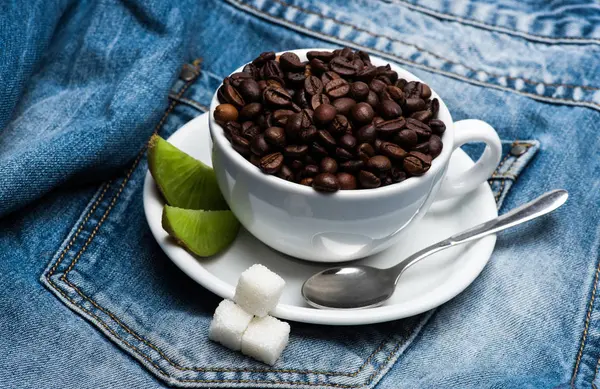 Kopp med kaffe bönor, raffinerat socker och sked på tallrik, denim bakgrund. Mugg full av kaffebönor på jeans. Nybryggt kaffe konceptet — Stockfoto