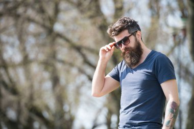 Hipster adam sokakta çıplak ağaçlar ile poz trendy güneş gözlüğü. Adam dövme onun kol yan görünümünde kentsel sahne üzerinde koyu mavi tişört içinde güneşli günde sakallı