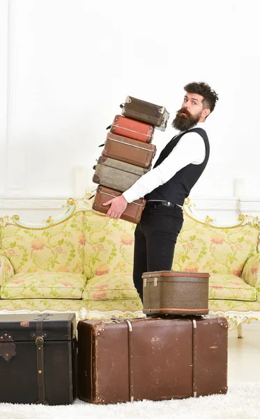 Man with beard and mustache wearing classic suit delivers luggage, luxury white interior background. Butler and service concept. Macho, elegant porter on strict face carries pile of vintage suitcases