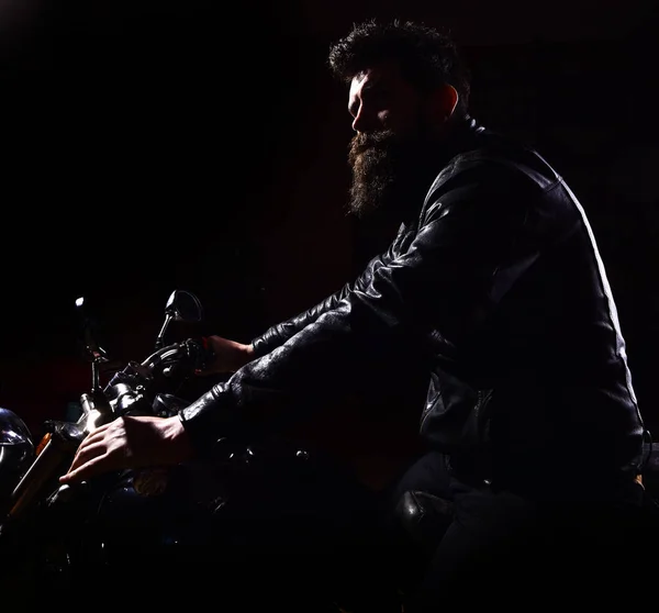 Hipster, motociclista brutale in giacca di pelle guida moto durante la notte, spazio per la copia. Concetto di cavaliere notturno. Uomo con barba, motociclista in giacca di pelle seduto sulla moto al buio, fondo nero — Foto Stock