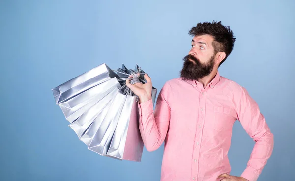 Sale and discount concept. Hipster on serious face carries shopping bags. Guy shopping on sales season with discounts. Man with beard and mustache works as shop assistant, light blue background