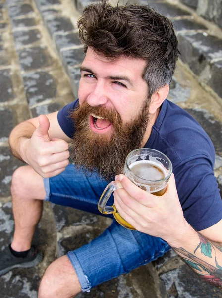 Homem com barba e bigode segura vidro com cerveja e mostra polegar para cima, fundo escadas de pedra. Hipster na cara alegre beber cerveja de esboço ao ar livre. Macho descansando com cerveja. Sexta-feira relaxar conceito — Fotografia de Stock