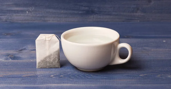 Concept d'heure du thé. tasse ou tasse en porcelaine blanche avec de l'eau chaude transparente et sac de thé. Processus de brassage du thé dans une tasse en céramique. Tasse remplie d'eau bouillante et sachet de thé sur fond de bois bleu — Photo
