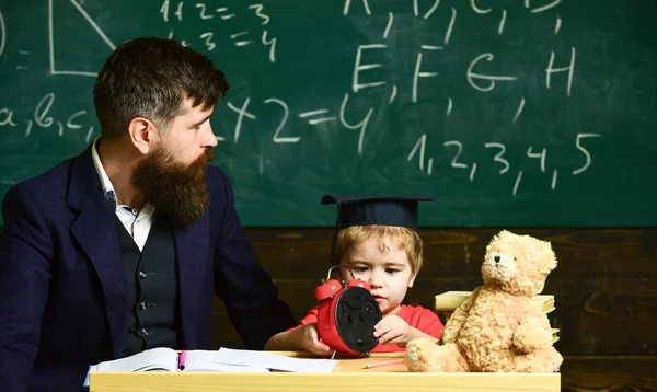 Teacher and pupil sitting in the classroom. Adult man in smart suit is looking to the side while kid is playing with clock. Small boy in mortarboard behind the table with pile of books and teddy bear