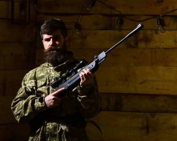 Man with beard wears camouflage clothing, wooden interior background. Macho on strict face at gamekeepers house. Huntsman concept. Hunter, brutal hipster with gun prepare rifle for hunting — Stock Photo, Image