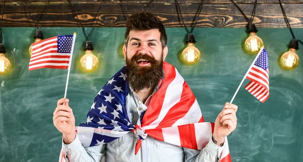 Patriottische opvoeding concept. Student exchange programma. Man met baard en snor op blij gezicht houdt vlaggen van de VS, in de klas, schoolbord op achtergrond. Amerikaanse leraar golven met Amerikaanse vlaggen — Stockfoto