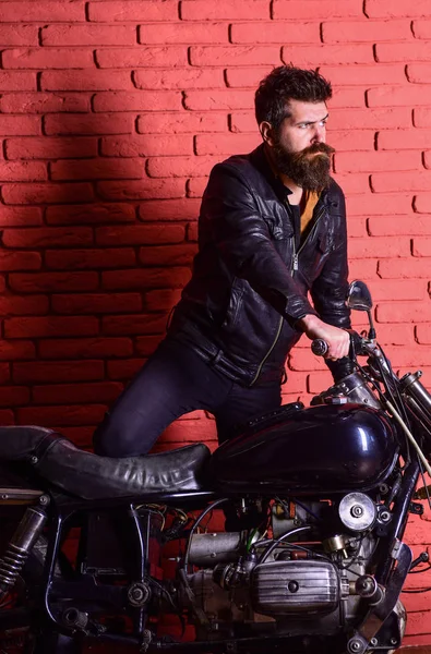 Хипстер, жестокий байкер на серьезном лице в кожаной куртке садится на мотоцикл. Мужская страсть. Мужчина с бородой, байкер в кожаной куртке возле мотоцикла в гараже, кирпичный фон стены — стоковое фото