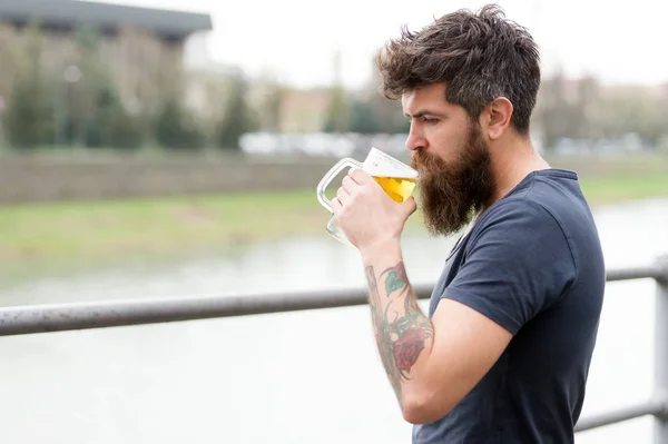 Hombre barbudo sostiene taza de cerveza, bebe cerveza al aire libre. El hombre con barba larga parece relajado. Hombre con barba y bigote en la cara tranquila, fondo del río, desenfocado. Concepto de cerveza artesanal — Foto de Stock