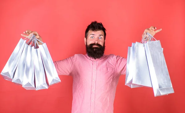 Bearded man shocked with amount of stuff bought on sale, shopping concept. Hipster with surprised face and long beard wearing pink shirt. Bearded man holding silver paper bags from fashionable store — Stock Photo, Image