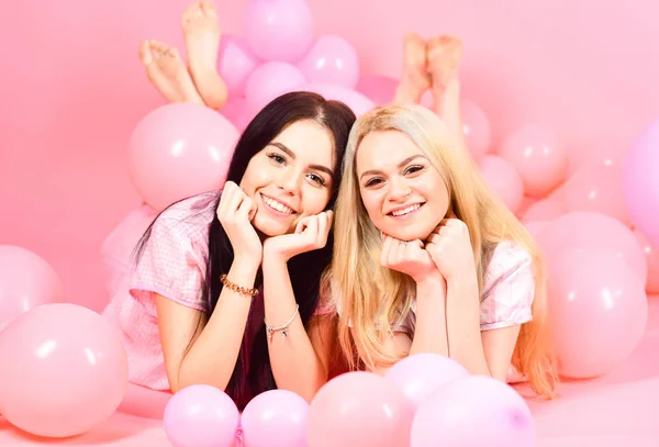 Brünette und blonde niedliche Frauen in rosa Pyjamas posieren vor der Kamera, während sie in der Nähe von Luftballons auf einer Schlummerparty vor rosa Hintergrund liegen. — Stockfoto