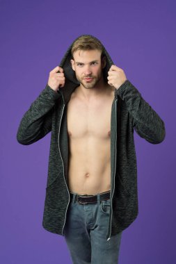 Gülen suratlı adam kapüşonlu, mor arka planlı ceketi deniyor. Yüzünde kıllar, göğsünde pürüzsüz bir deri olan adam giyinmeye çalışıyor. Moda konsepti. Adam uzun kapüşonlu ceketle çekici görünüyor.