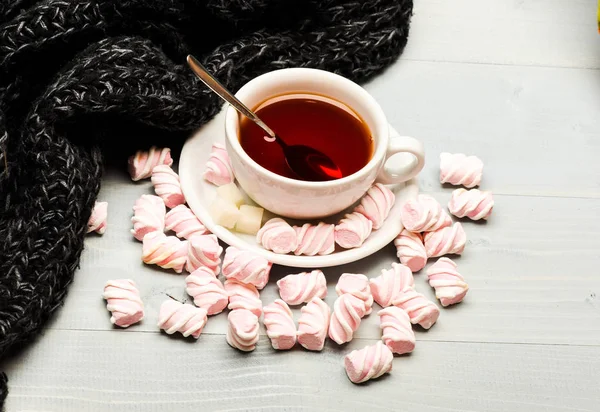 Mug and sweets near cozy knitted scarf. Mug filled with black brewed tea, spoon and heap of marshmallow on white wooden background. Cup with hot black tea and marshmallows. Coziness concept — Stock Photo, Image