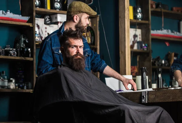 Homme à la barbe recouverte d'une cape noire attendant pendant que le coiffeur change de grade de tondeuse. Un client hippie qui se fait couper les cheveux. Client avec barbe prête pour la coupe ou le toilettage. Concept de processus de coupe de cheveux — Photo