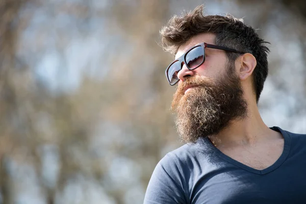 Il tizio con la barba porta gli occhiali da sole. Hipster con barba e baffi sul viso rigoroso, sfondo naturale, sfocato. L'uomo con la barba sembra elegante e fiducioso nella giornata di sole. Moda e stile concetto — Foto Stock